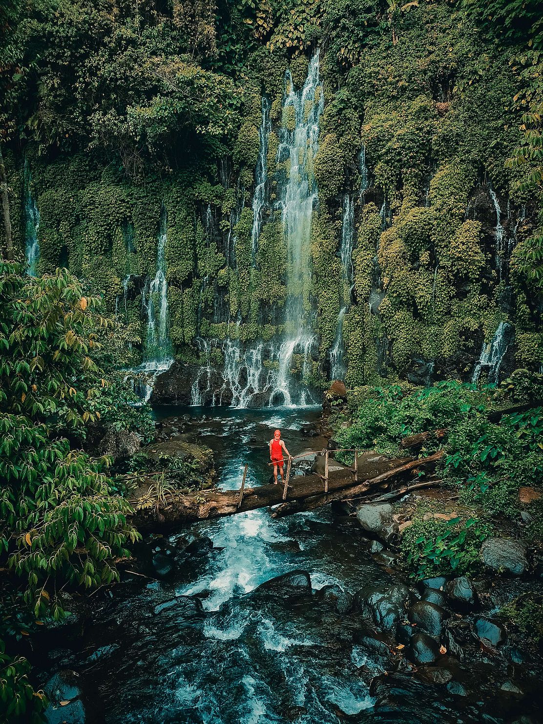 Auf diesem Bild sieht man, wie ein Mann auf einer Brücke vor einem Wasserfall im Dschungel steht. 