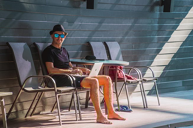 Ein Mann sitzt auf einem Stuhl und hat seinen Lauptop auf dem Schoß. Er hat einen Hut auf und eine Sonnenbrille. Er lächelt in die Kamera.