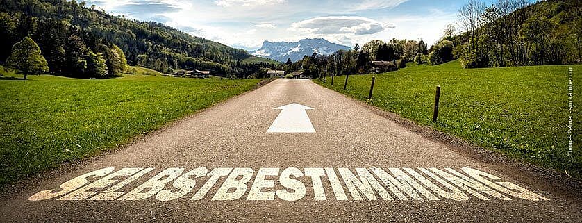Auf diesem Bild ist eine Straße in der Natur zu sehen, mit der Aufschrift "Selbstbestimmung" und einen Pfeil nach vorne